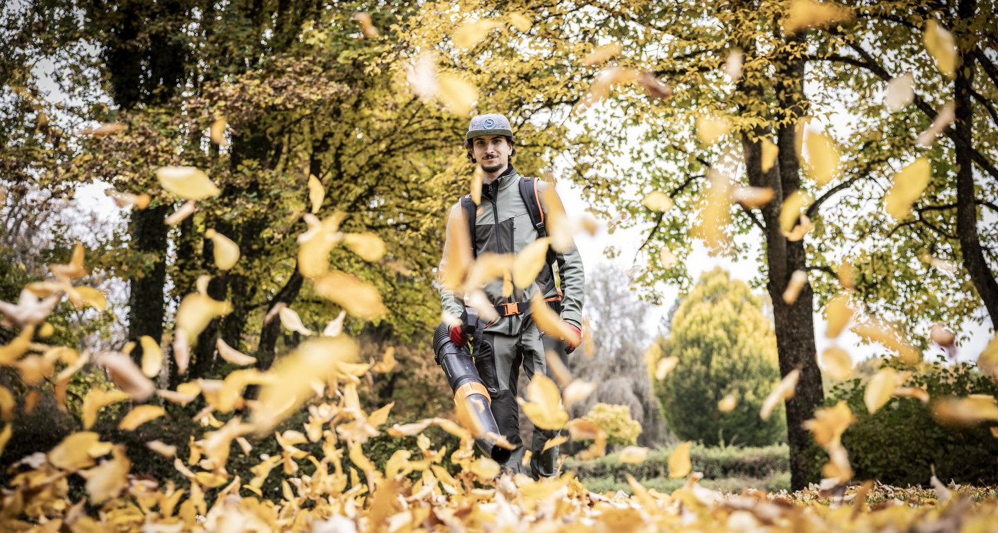 Mit einem starken Akkulaubbläser wird Laub, das im Herbst gefallen ist in die Luft geblasen. Im Hintergrund erkennt man den Mitarbeiter zwischen Bäumen.