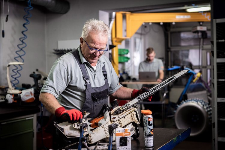 Erfahrener Mechanikermeister repariert eine Stihl Motorsäge. Im Hintergrund sieht man einen weiteren Mitarbeiter, der mit dem Laptop einen Profi Balkenmäher der Firma Brielmaier ausliest.