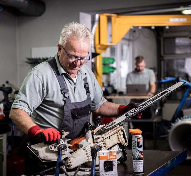 Erfahrener Mechanikermeister repariert eine Stihl Motorsäge. Im Hintergrund sieht man einen weiteren Mitarbeiter, der mit dem Laptop einen Profi Balkenmäher der Firma Brielmaier ausliest.