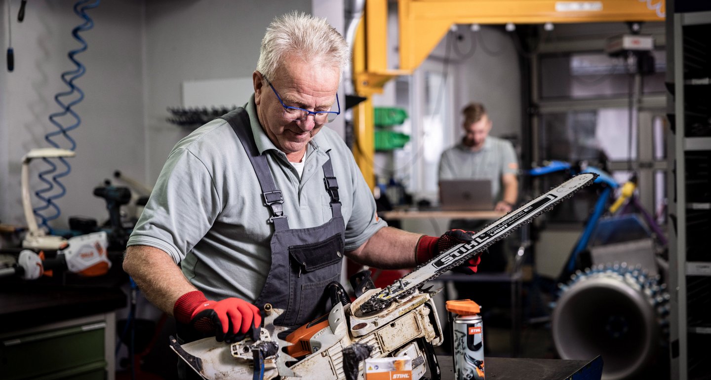 Erfahrener Mechanikermeister repariert eine Stihl Motorsäge. Im Hintergrund sieht man einen weiteren Mitarbeiter, der mit dem Laptop einen Profi Balkenmäher der Firma Brielmaier ausliest.