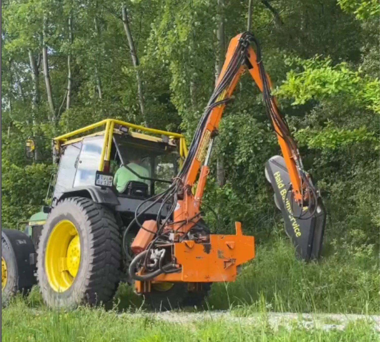 Ein Traktor mit einem langen, orangefarbenen Mäharm mäht Gras und Büsche am Straßenrand vor einem Wald mit grünen Bäumen.