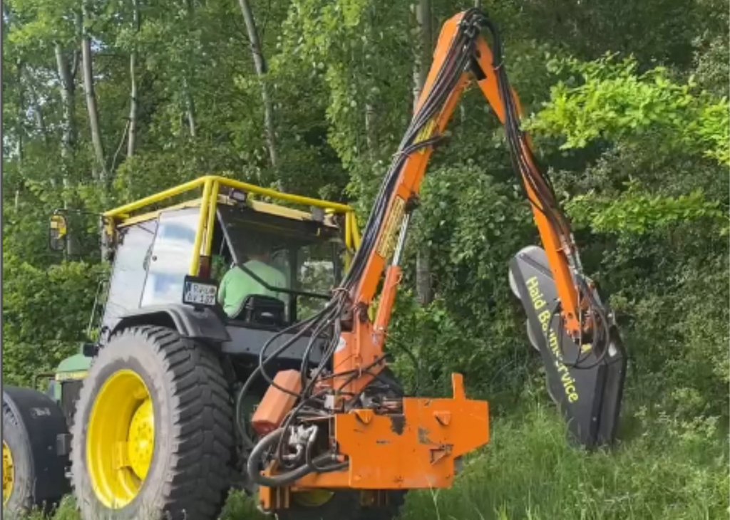 Ein Traktor mit einem langen, orangefarbenen Mäharm mäht Gras und Büsche am Straßenrand vor einem Wald mit grünen Bäumen.