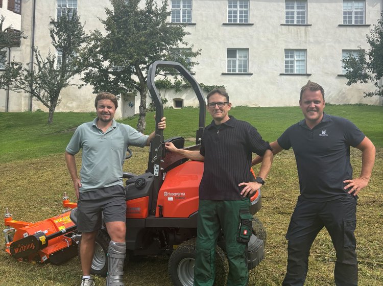 Drei Männer stehen lachend zusammen auf einer Rasenfläche vor einem historischen Gebäude. Einer der Männer lehnt an einem orangefarbenen Aufsitzmäher. Alle wirken entspannt und freundlich.