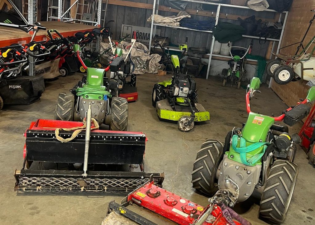 Mehrere motorbetriebene Garten- und Grünflächenmäher stehen in einer Halle, teils mit großen Rädern und Bürstenaufbauten zur Rasenpflege. Regale mit weiterem Gerät sind im Hintergrund sichtbar.