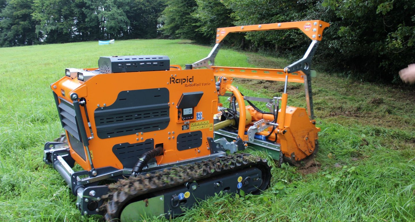 Ein oranger, kettengetriebener Mulchroboter arbeitet auf einer Wiese und zerkleinert Gras und Pflanzenrückstände.