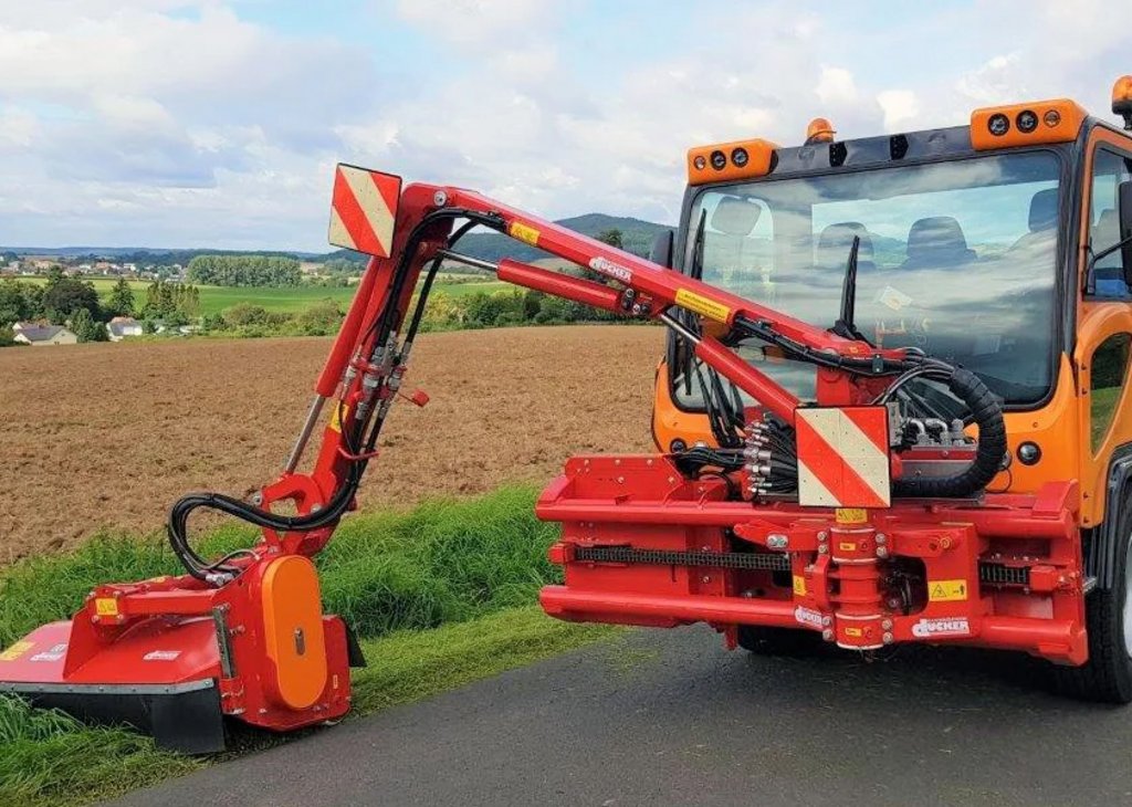 Das Bild zeigt einen Transporter der Firma Ladog. Er hat ein Mähwerk angebaut und mäht den Straßenrand neben einem Feld.
