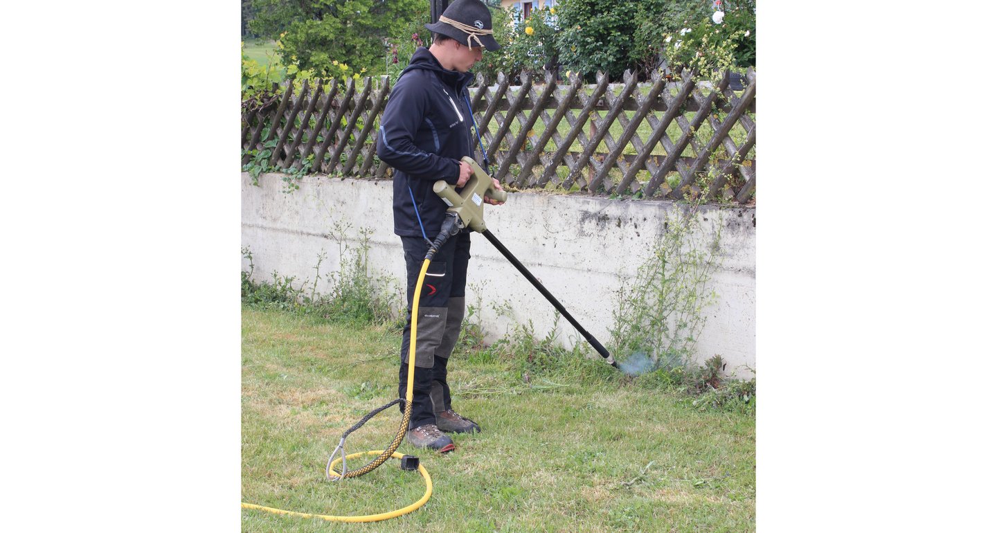 Ein Mann steht vor einer Gartenmauer. Er hält eine Elektrolanze in der Hand zum Unkraut vernichten.