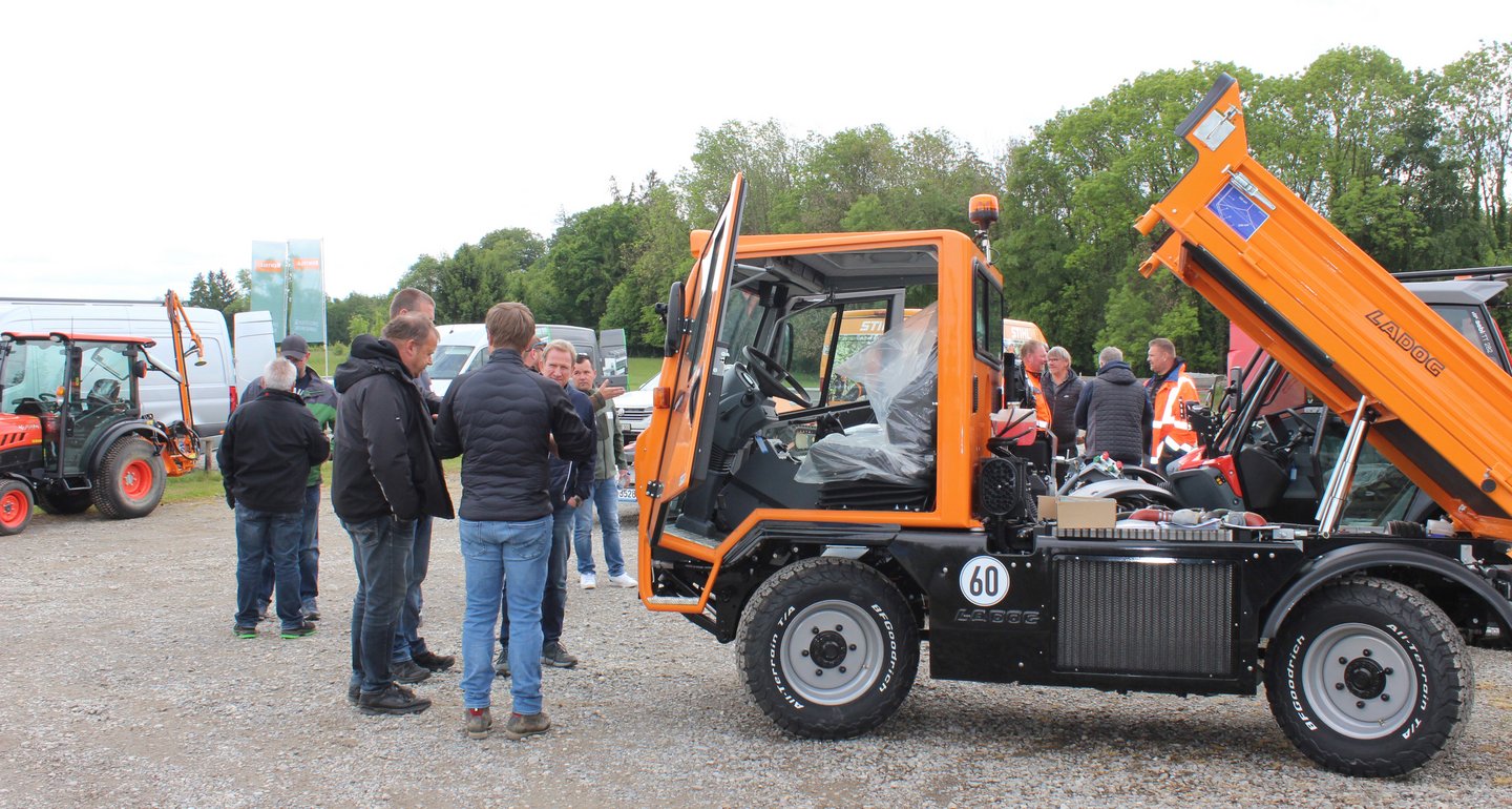 Auf einem Kiesplatz steht ein Mehrzwecktransporter im Vordergrund. Eine Gruppe von Männern stehen um das Fahrzeug und besichtigen es.