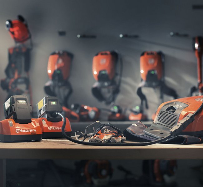 Auf einem Tisch stehen zwei Akku-Ladegeräte, ein Laubbläser und Arbeitshandschuhe, im Hintergrund sind verschiedene orangefarbene Motorsägen und Schutzkleidung an der Wand aufgehängt. Alle Geräte sind von der Marke Husqvarna.