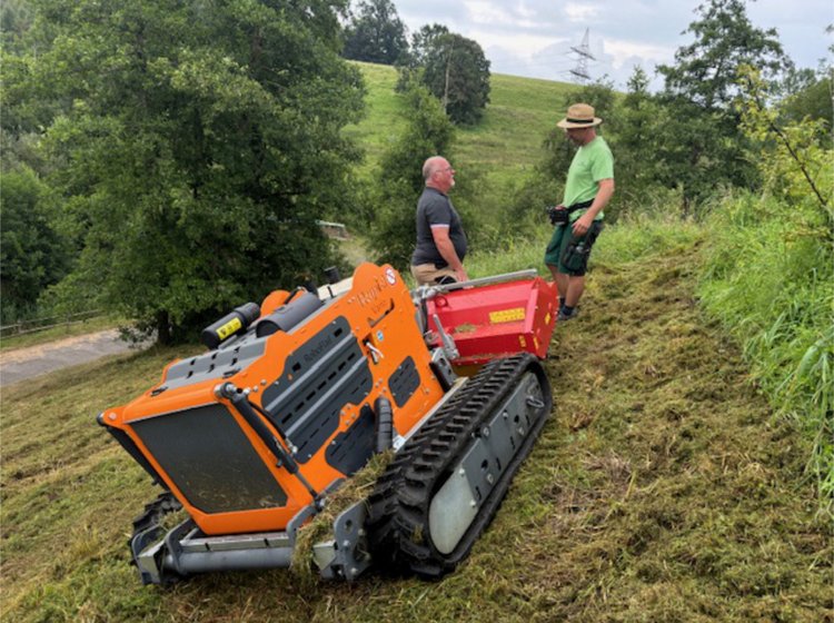 Zwei Männer bedienen eine orange Raupendrohne auf einem grasbewachsenen Hügel, um Vegetation zu mähen.