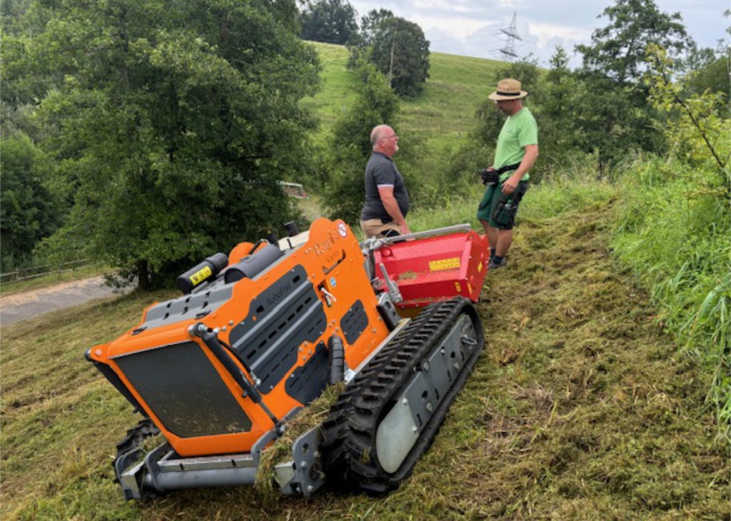 Zwei Männer bedienen eine orange Raupendrohne auf einem grasbewachsenen Hügel, um Vegetation zu mähen.