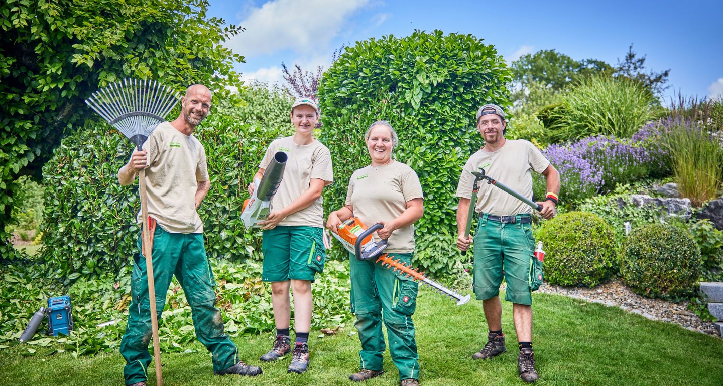 Vier Gärtner stehen in einem Garten und halten verschiedene Gartengeräte wie Rechen, Laubbläser und Heckenschere. Sie tragen Arbeitskleidung und lächeln in die Kamera. Der Hintergrund zeigt gepflegte Hecken, Pflanzen und einen blauen Himmel.