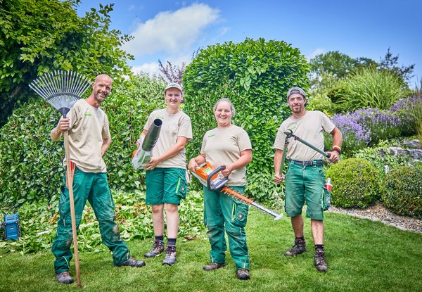 Vier Gärtner stehen in einem Garten und halten verschiedene Gartengeräte wie Rechen, Laubbläser und Heckenschere. Sie tragen Arbeitskleidung und lächeln in die Kamera. Der Hintergrund zeigt gepflegte Hecken, Pflanzen und einen blauen Himmel.