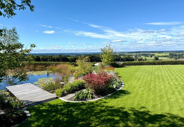 Ein weitläufiges Grundstück bei Sonnenschein. Am Horizont sieht man weiße Wolken am Himmel. Im Vordergrund ist ein großes Beet mit Sträuchern und Blumen. Ein Holzsteg führt in einen Teich mit Seerosen.