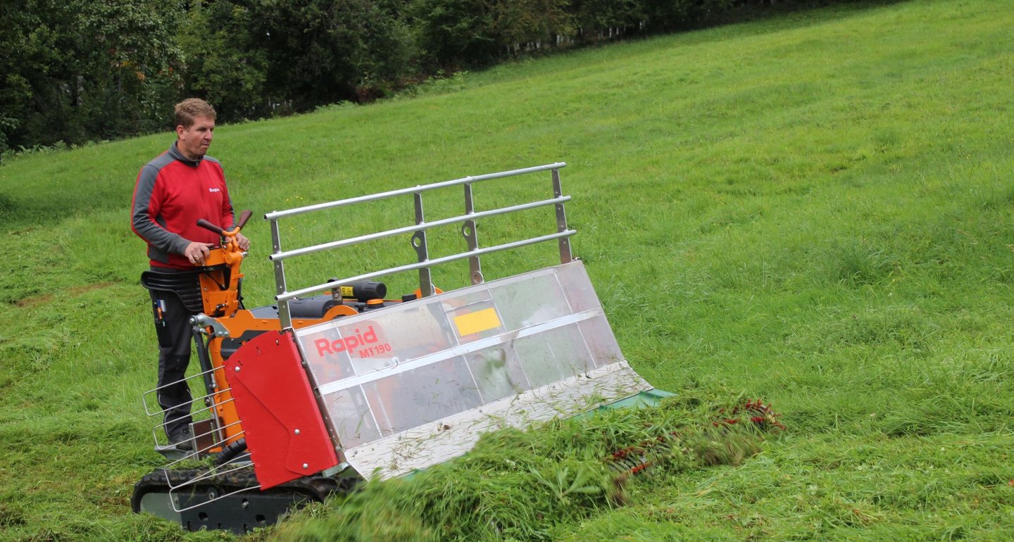 Ein Mann steuert eine kleine ferngesteuerte Baumaschine, die Gras auf einer Wiese mäht.