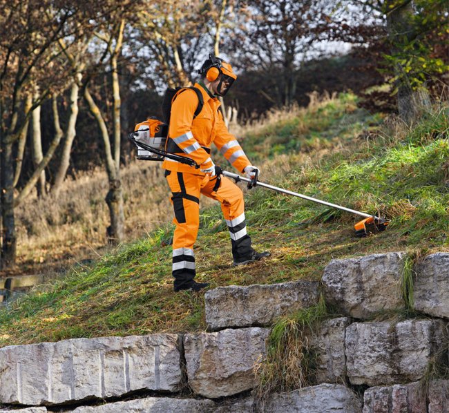 Ein Arbeiter in orangefarbener Schutzkleidung mäht Gras an einem Hang mit einem Freischneider.