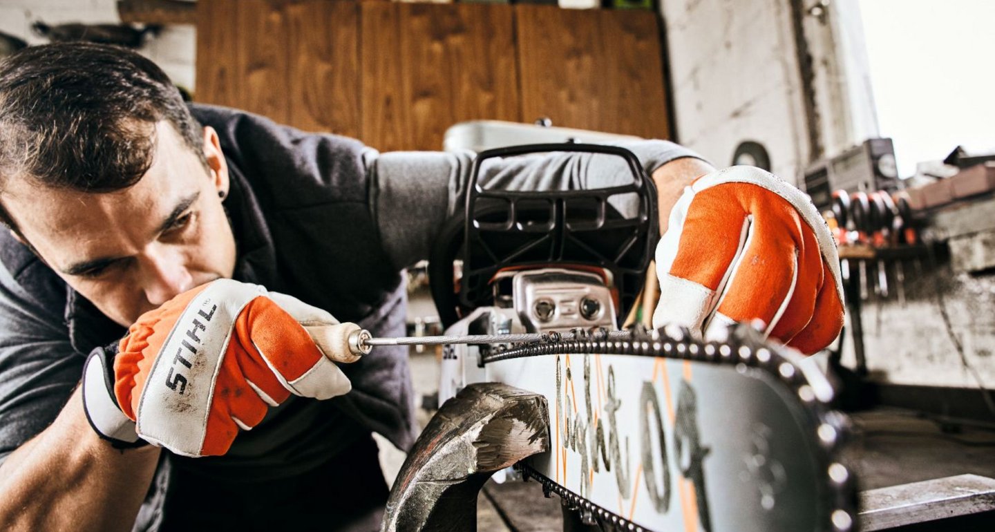 Ein Mann überprüft und schärft sorgfältig die Kette einer Motorsäge in einer Werkstatt. Er trägt Schutzhandschuhe und konzentriert sich auf die genaue Arbeit an der Sägekette.