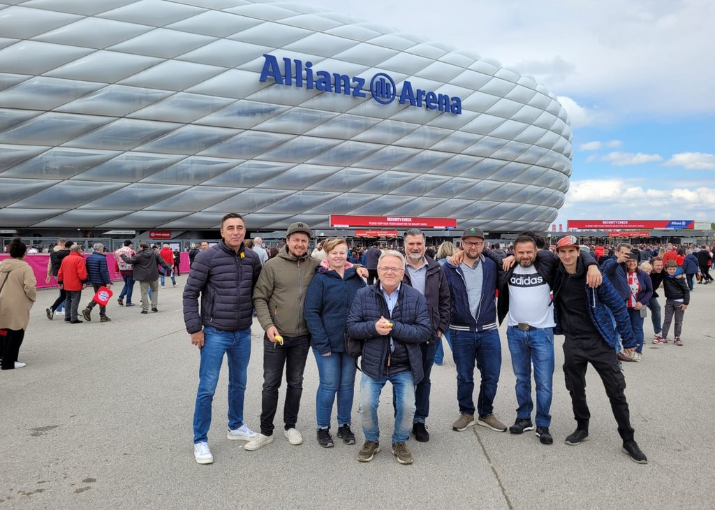 Gruppe von acht Personen steht vor der markanten, gebogenen Außenfassade der Allianz Arena, einem Fußballstadion in München, mit vielen weiteren Menschen im Hintergrund.