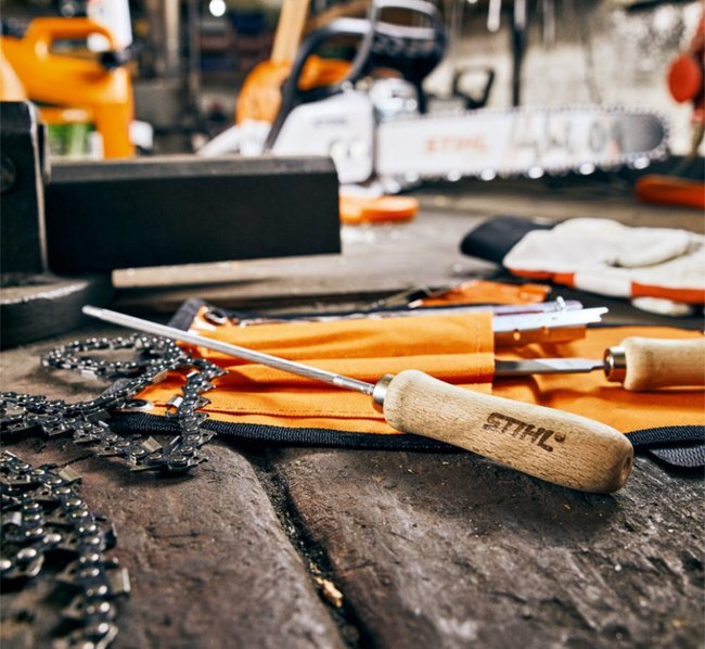 Werkstatt mit Kettensäge-Wartungswerkzeugen auf einem Holztisch, darunter eine Kettensägekette, Schraubenzieher mit Holzgriff und orangefarbene Schutzhandschuhe. Im Hintergrund weitere Werkzeuge und Maschinen unscharf erkennbar.