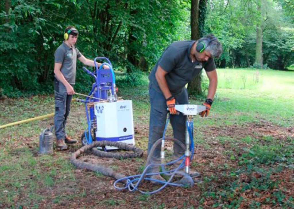 Zwei Personen tragen Gehörschutz und bedienen im Wald eine Maschine, die mit Schläuchen verbunden ist, um den Boden zu bearbeiten oder Proben zu entnehmen.