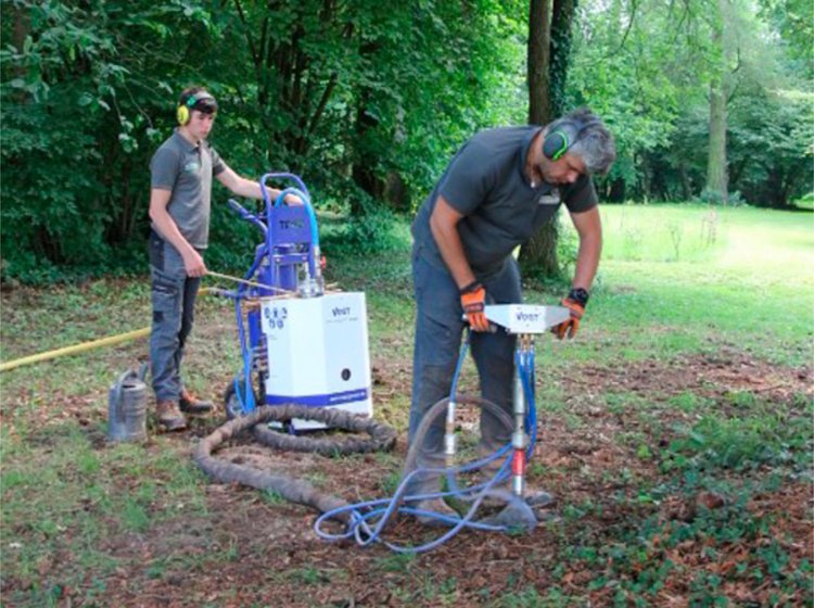 Zwei Personen tragen Gehörschutz und bedienen im Wald eine Maschine, die mit Schläuchen verbunden ist, um den Boden zu bearbeiten oder Proben zu entnehmen.
