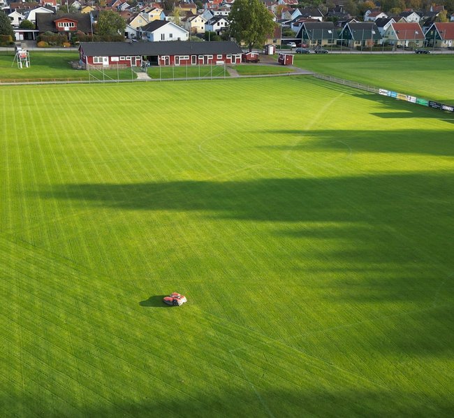 Ein großer, gepflegter Fußballrasen mit Markierungen für ein Spielfeld, umgeben von Tribünen und Häusern im Hintergrund. Ein Rasenmähroboter fährt auf dem Spielfeld.