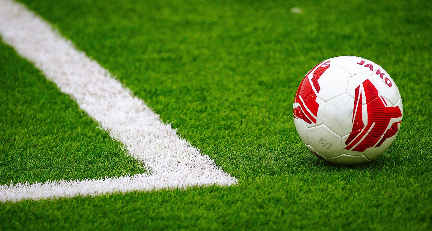 Auf dem Foto ist ein kleiner Ausschnitt eines Fußballplatzes zu sehen: Grüner Rasen, eine Markierungsecke und davor ein rot-weißer Fußball.