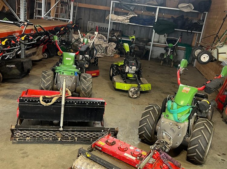 Mehrere motorbetriebene Garten- und Grünflächenmäher stehen in einer Halle, teils mit großen Rädern und Bürstenaufbauten zur Rasenpflege. Regale mit weiterem Gerät sind im Hintergrund sichtbar.