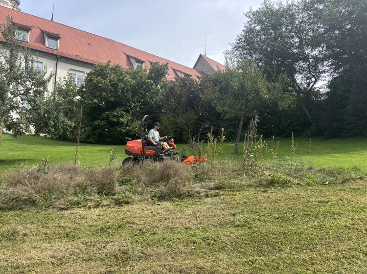 Mann fährt mit einem Aufsitzmäher und mäht eine große Grünfläche vor einem Gebäude mit rotem Dach.
