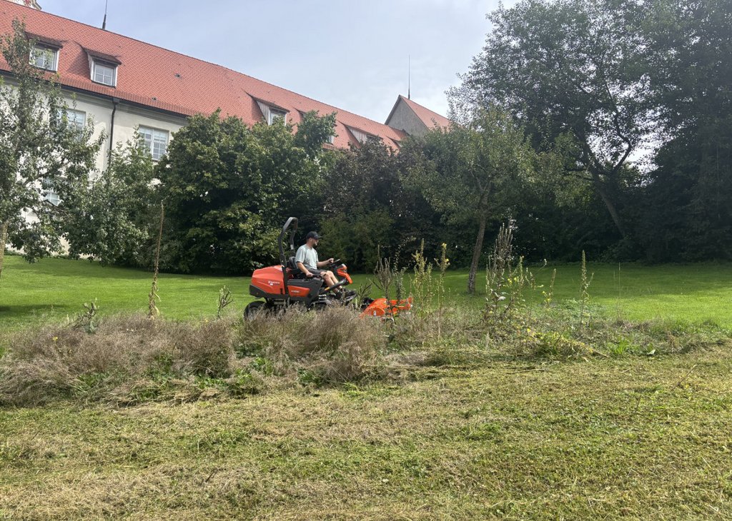 Mann fährt mit einem Aufsitzmäher und mäht eine große Grünfläche vor einem Gebäude mit rotem Dach.
