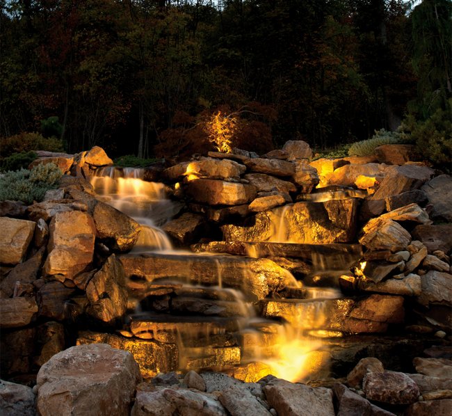 Beleuchteter, mehrstufiger Wasserfall aus Felsen in einem Garten, umgeben von Bäumen, aufgenommen in der Dämmerung.