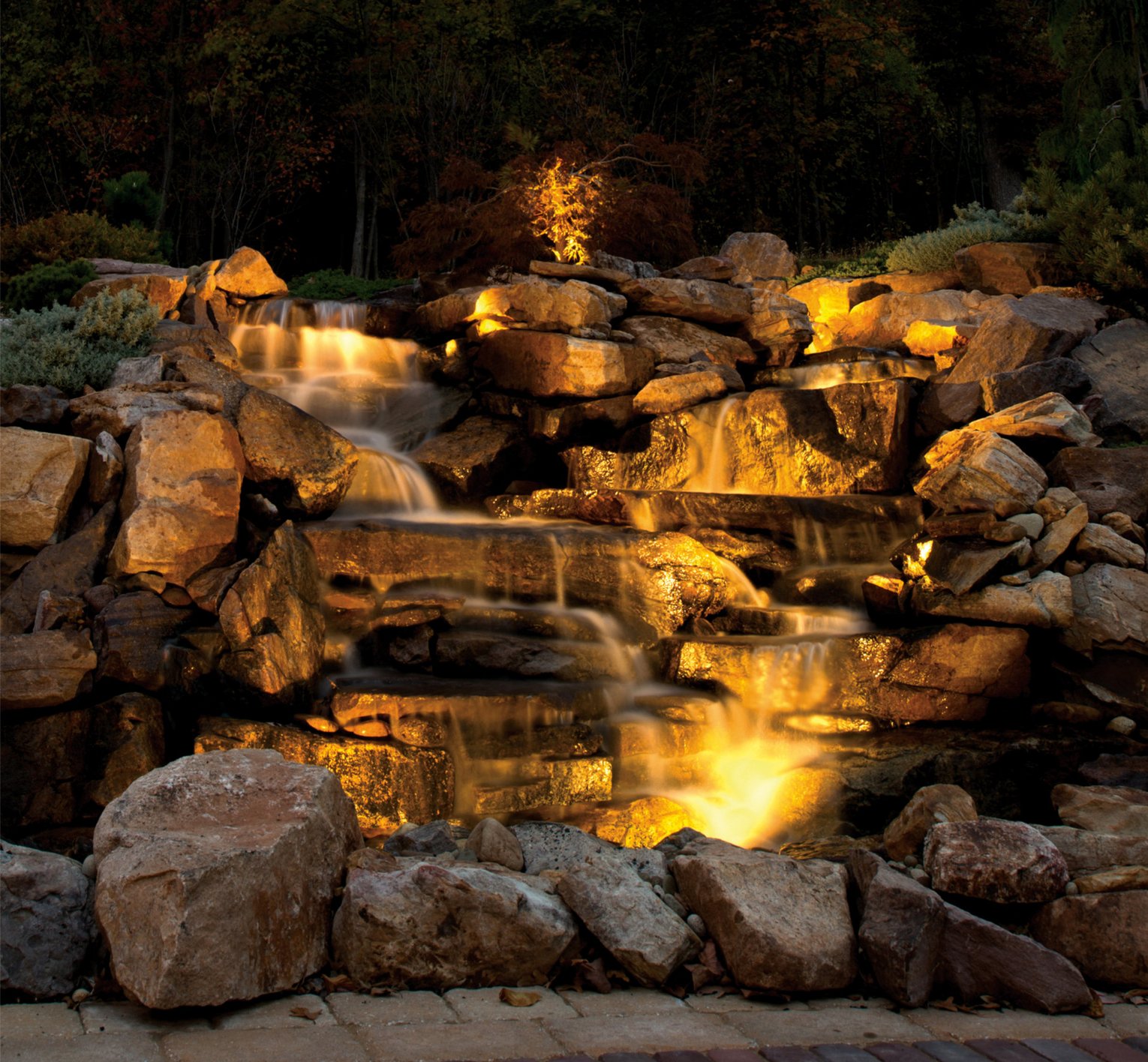 Beleuchteter, mehrstufiger Wasserfall aus Felsen in einem Garten, umgeben von Bäumen, aufgenommen in der Dämmerung.