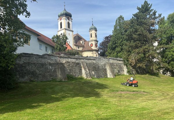 Historische Stadtmauer im Vordergrund, im Hintergrund zwei barocke Kirchtürme mit Zwiebelkuppeln, umgeben von Bäumen. Auf der Wiese mäht eine Person mit einem Rasenmäher das Gras.
