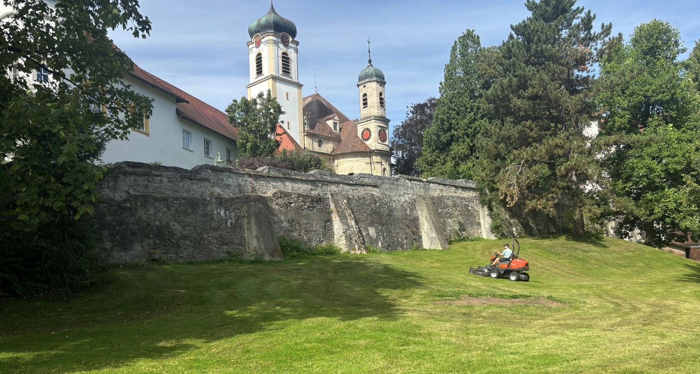 Historische Stadtmauer im Vordergrund, im Hintergrund zwei barocke Kirchtürme mit Zwiebelkuppeln, umgeben von Bäumen. Auf der Wiese mäht eine Person mit einem Rasenmäher das Gras.