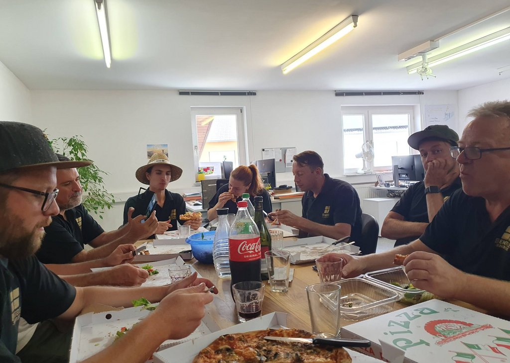 Eine Gruppe von sechs Erwachsenen sitzt um einen Tisch, isst Pizza und trinkt Getränke in einem hellen Raum, der wie ein Büro wirkt. Auf dem Tisch steht eine große Flasche Coca-Cola, und alle wirken entspannt und in Gesellschaft.