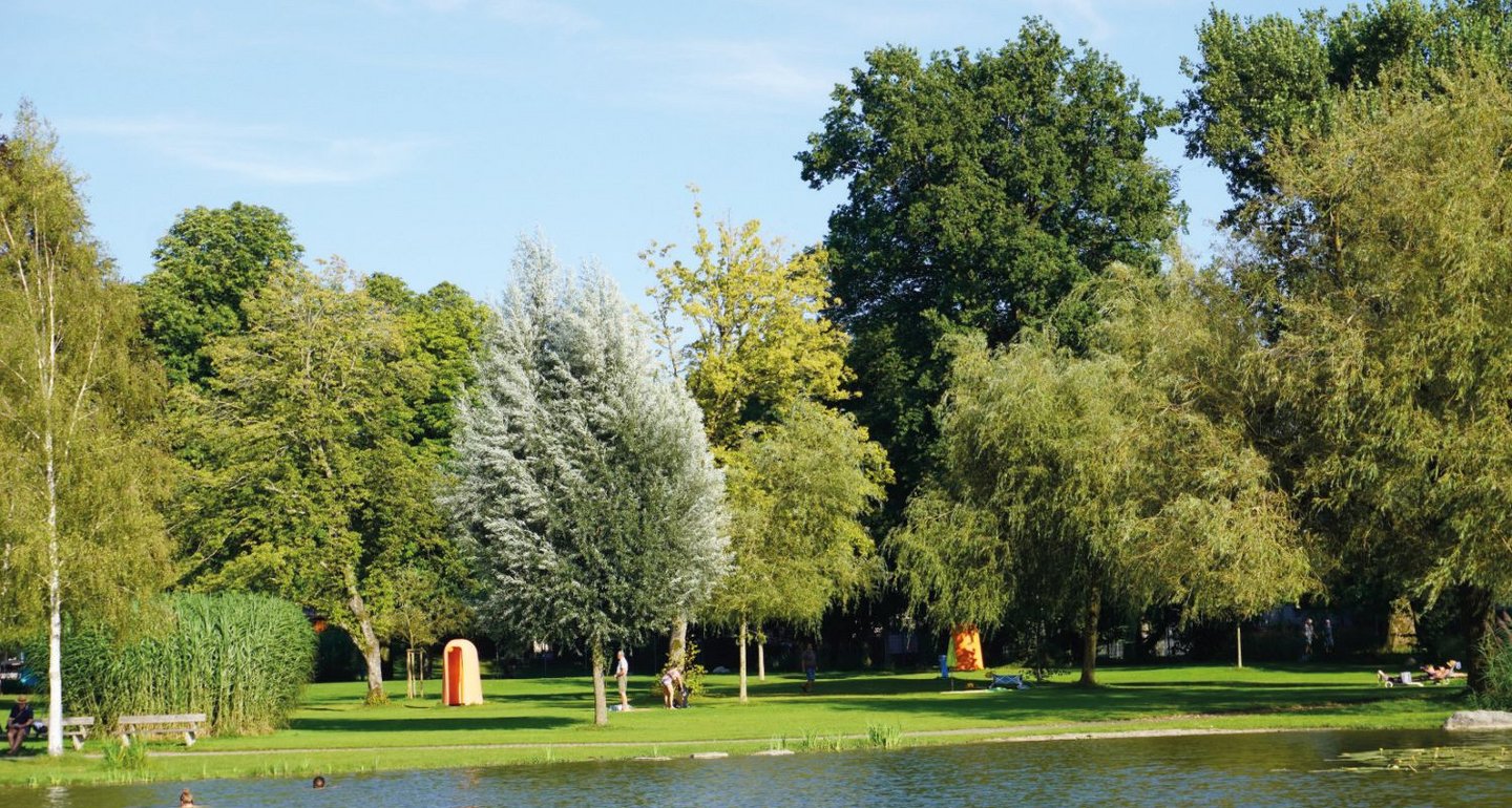 Ein ruhiger Park mit einem Teich im Vordergrund, umgeben von verschiedenen grünen Bäumen unter klarem, blauem Himmel. Menschen sitzen entspannt auf der Wiese.