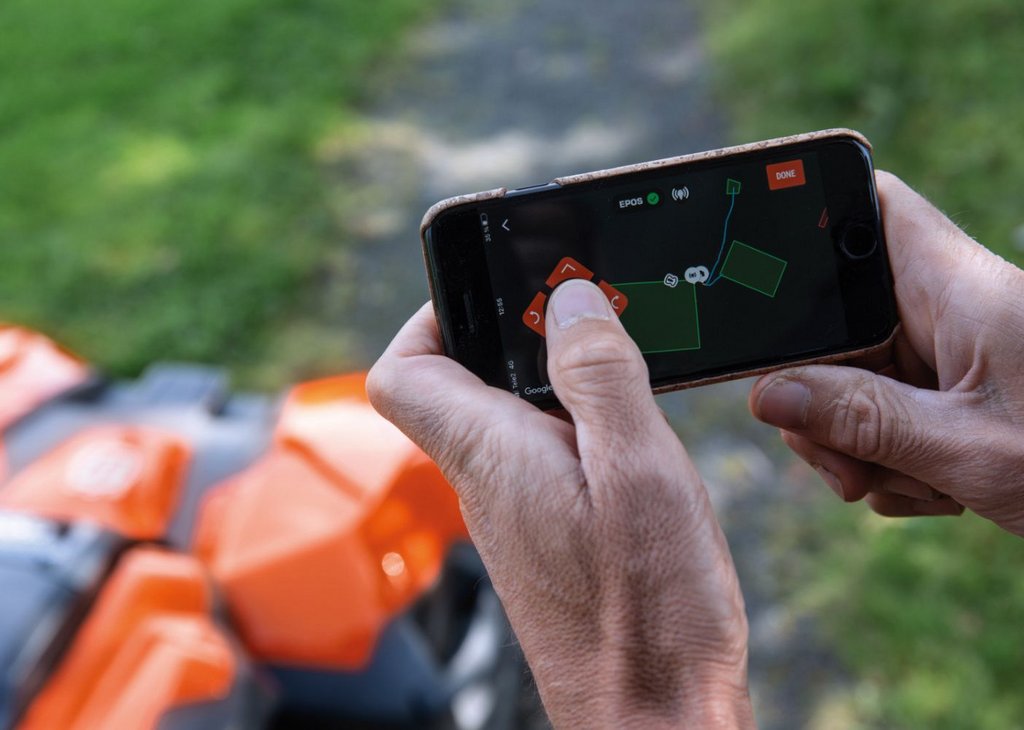 Eine Person steuert über eine App auf einem Smartphone einen orangefarbenen Mähroboter im Freien, sichtbar ist eine Kartenansicht der Mähfläche auf dem Bildschirm.