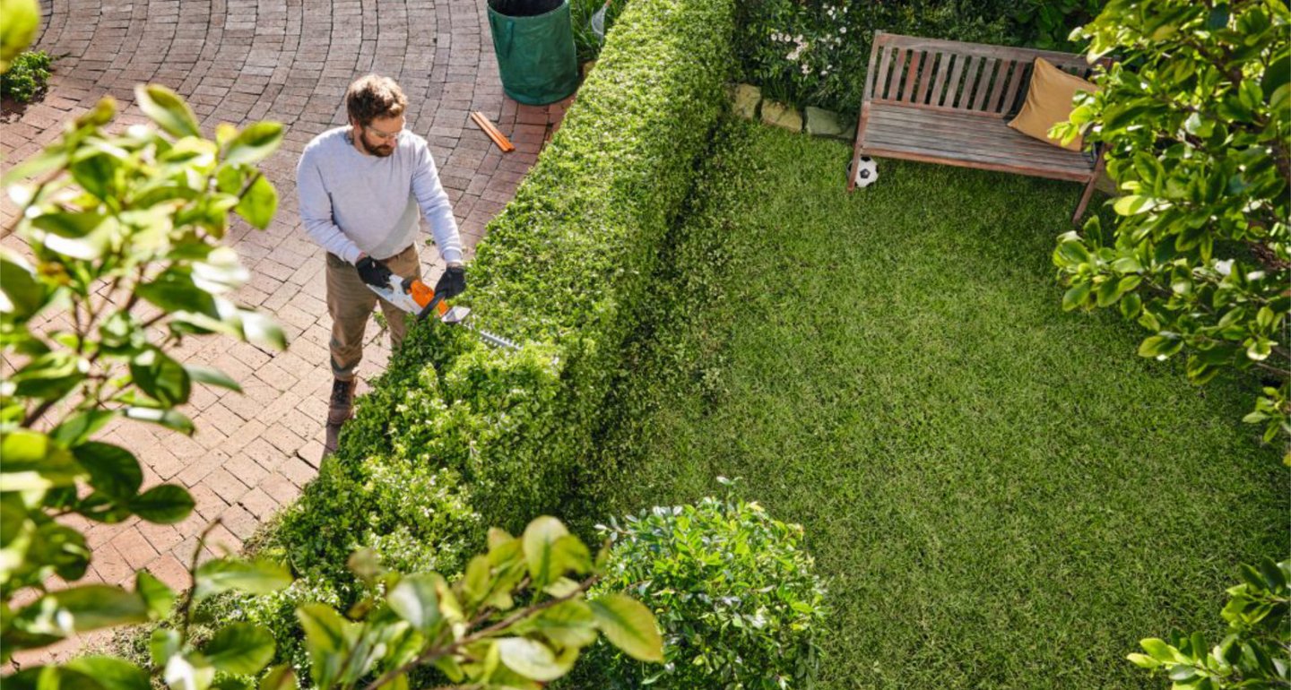Ein Mann schneidet mit einer Heckenschere eine grüne Gartenhecke in einem gepflegten Garten. Umgeben von Rasen, Büschen und einer Gartenbank mit einem Kissen.