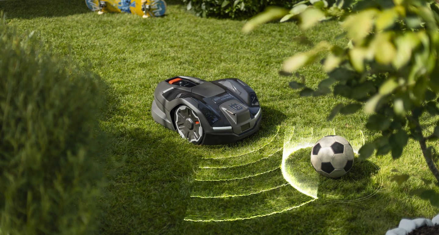Ein schwarzer Mähroboter mäht automatisch den Rasen und erkennt dabei mithilfe eines Sensors einen abgestellten Fußball, um Kollisionen zu vermeiden.