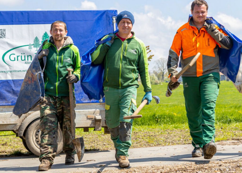 Drei Gartenarbeiter in Arbeitskleidung tragen Werkzeuge und Säcke, sie gehen fröhlich auf einer Wiese entlang, im Hintergrund ein Anhänger mit Firmenlogo.