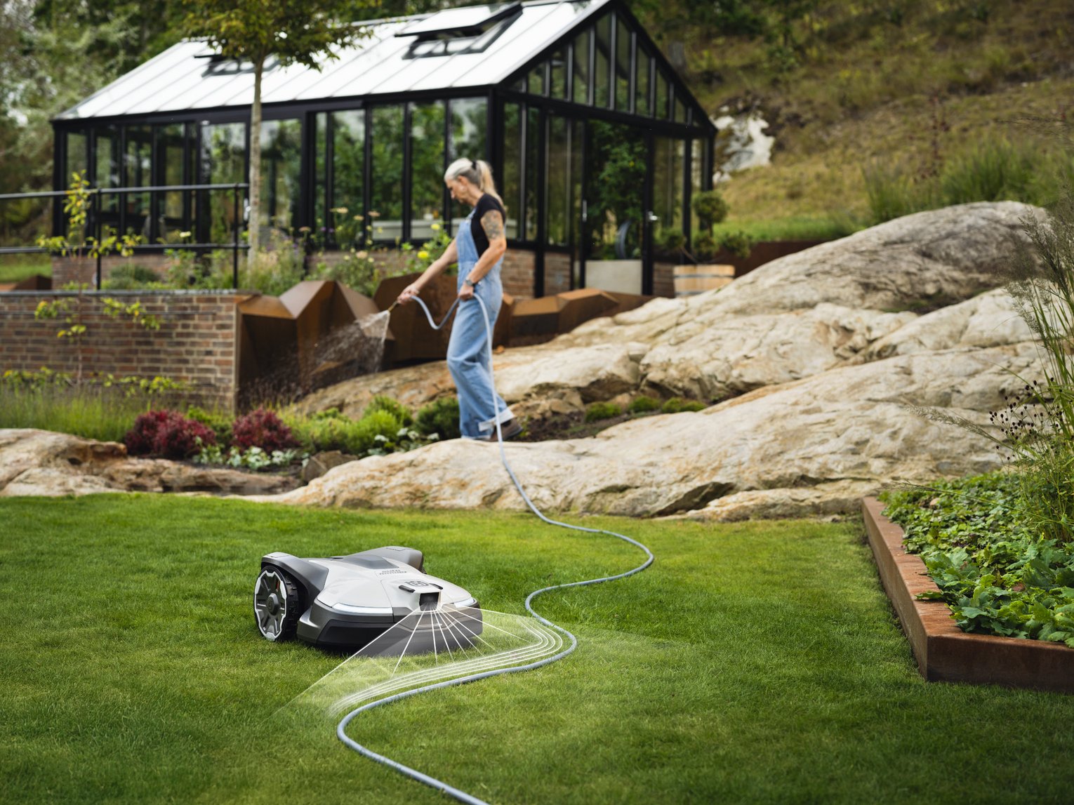 Eine Frau hält einen Gartenschlauch, während ein autonomer Rasenmähroboter den Rasen vor ihr mäht. Der Roboter navigiert selbstständig. Im Hintergrund ist ein Gewächshaus und eine Felsenlandschaft zu sehen.