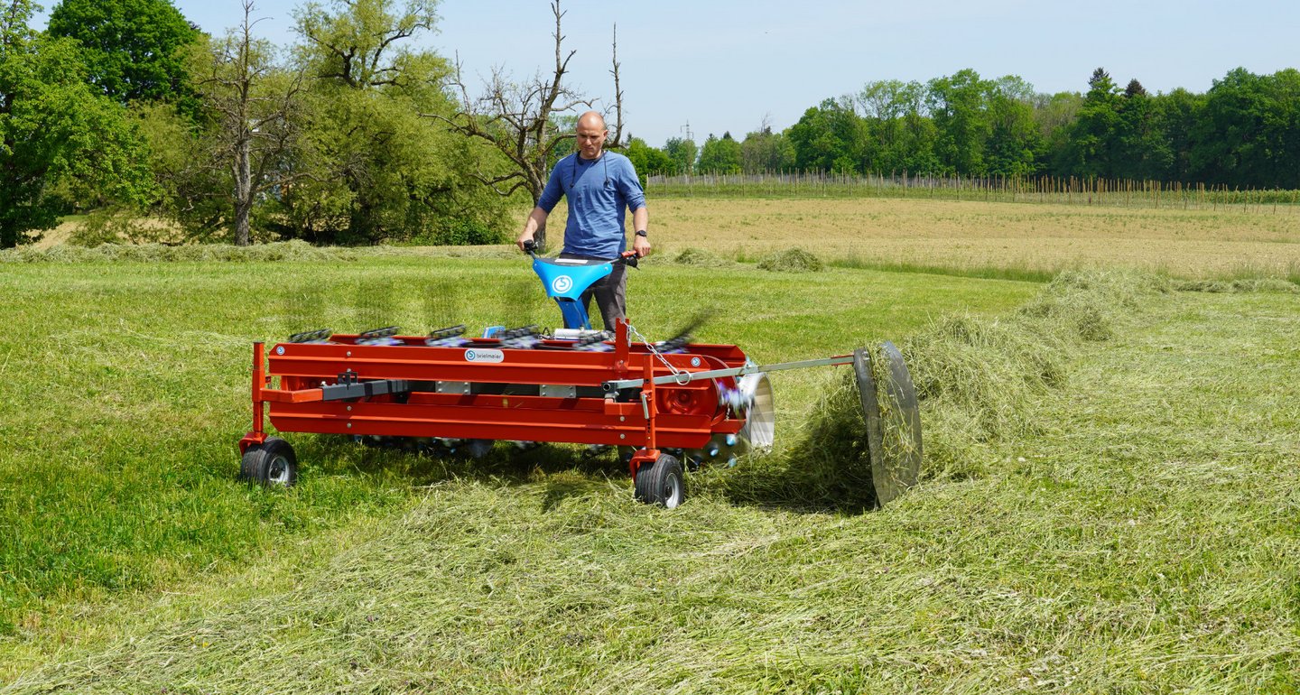 Ein Mann bedient eine rot lackierte landwirtschaftliche Maschine auf einer Wiese, die Grasschnitt zu einer Reihe zusammenlegt. Im Hintergrund sind Bäume und ein hellblauer Himmel zu sehen.