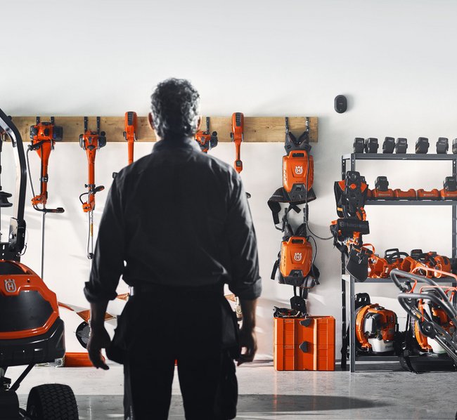 Ein Mann steht in einer Werkstatt vor einer Wand, an der diverse orangefarbene Gartengeräte und Werkzeuge ordentlich hängen und auf Regalen stehen.