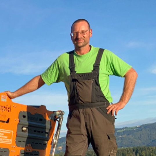 Ein Mann in Arbeitskleidung steht auf einer Wiese neben einem orangefarbenen Mulchgerät, das Gras und Vegetation mäht. Die Umgebung ist ländlich mit blauem Himmel und Hügeln im Hintergrund.