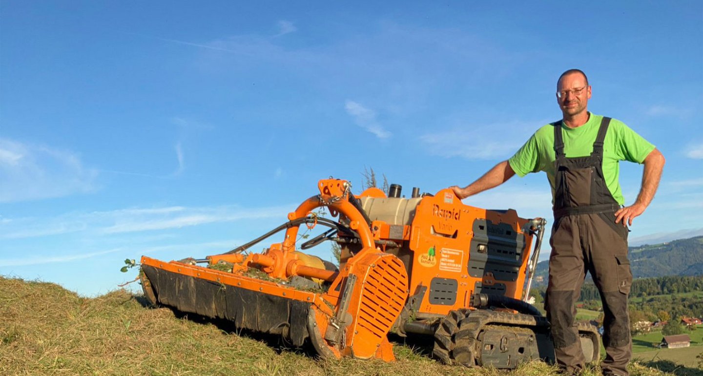 Ein Mann in Arbeitskleidung steht auf einer Wiese neben einem orangefarbenen Mulchgerät, das Gras und Vegetation mäht. Die Umgebung ist ländlich mit blauem Himmel und Hügeln im Hintergrund.