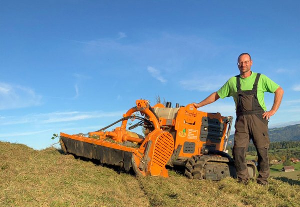 Ein Mann in Arbeitskleidung steht auf einer Wiese neben einem orangefarbenen Mulchgerät, das Gras und Vegetation mäht. Die Umgebung ist ländlich mit blauem Himmel und Hügeln im Hintergrund.
