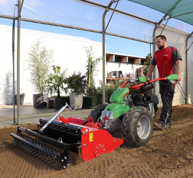 Ein Gartenarbeiter fräst den Boden eines Beets in einem Gewächshaus mit einer Umkehrfräse. Im Hintergrund sind Topfpflanzen.