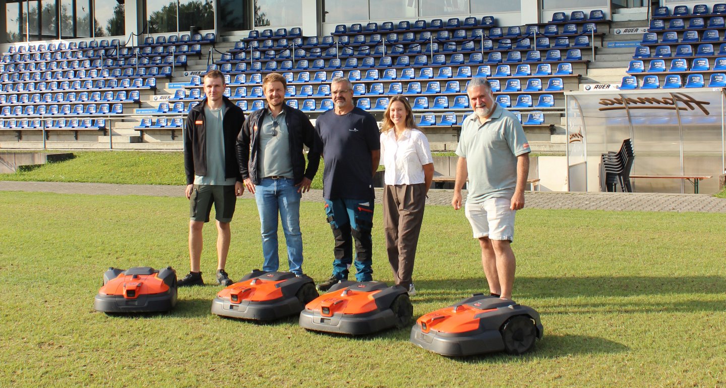 Im Fußballstadion der Stadt Ravensburg stehen vor der Tribüne David Meschenmoser (Fa. Bentele), Alexander Bentele (Fa. Bentele), Markus Reichle (Platzwart Stadt Ravensburg), Verena Riek (Stadt Ravensburg) und Ekrem Ozan (Fa. Bentele). Vor ihnen stehen vier Mähroboter.