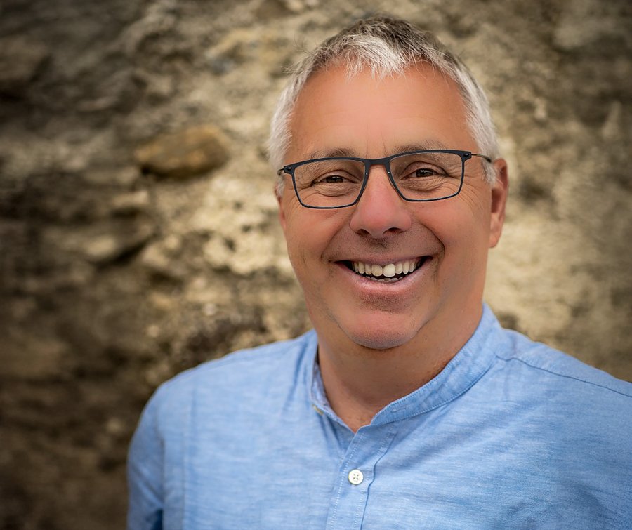 Porträt eines lächelnden Mannes mit grauen Haaren und Brille, der ein hellblaues Hemd trägt.