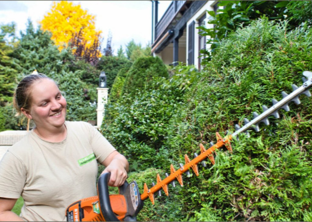 Frau trimmt mit elektrischem Heckenscheren-Gerät eine dichte grüne Hecke im Garten. Sie lächelt dabei.
